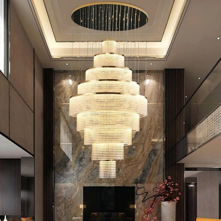 The Alleri Foyer Chandelier from Morsale.com, adorned with crystal embellishments, hangs elegantly from a high ceiling in a modern, opulent interior. This luxurious chandelier illuminates the marble wall and sleek dark wood panels, creating an extravagant ambiance. A glass balcony is visible on the right.