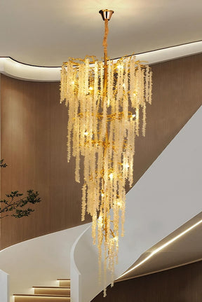 The Albero Duplex Staircase Foyer Crystal Chandelier from Morsale.com, featuring luxurious cascading beaded strands and polished gold accents, hangs elegantly from the ceiling, warmly illuminated. It is set against a modern interior with curved wooden walls and a white spiraling staircase in the background. A small plant is visible to the left.