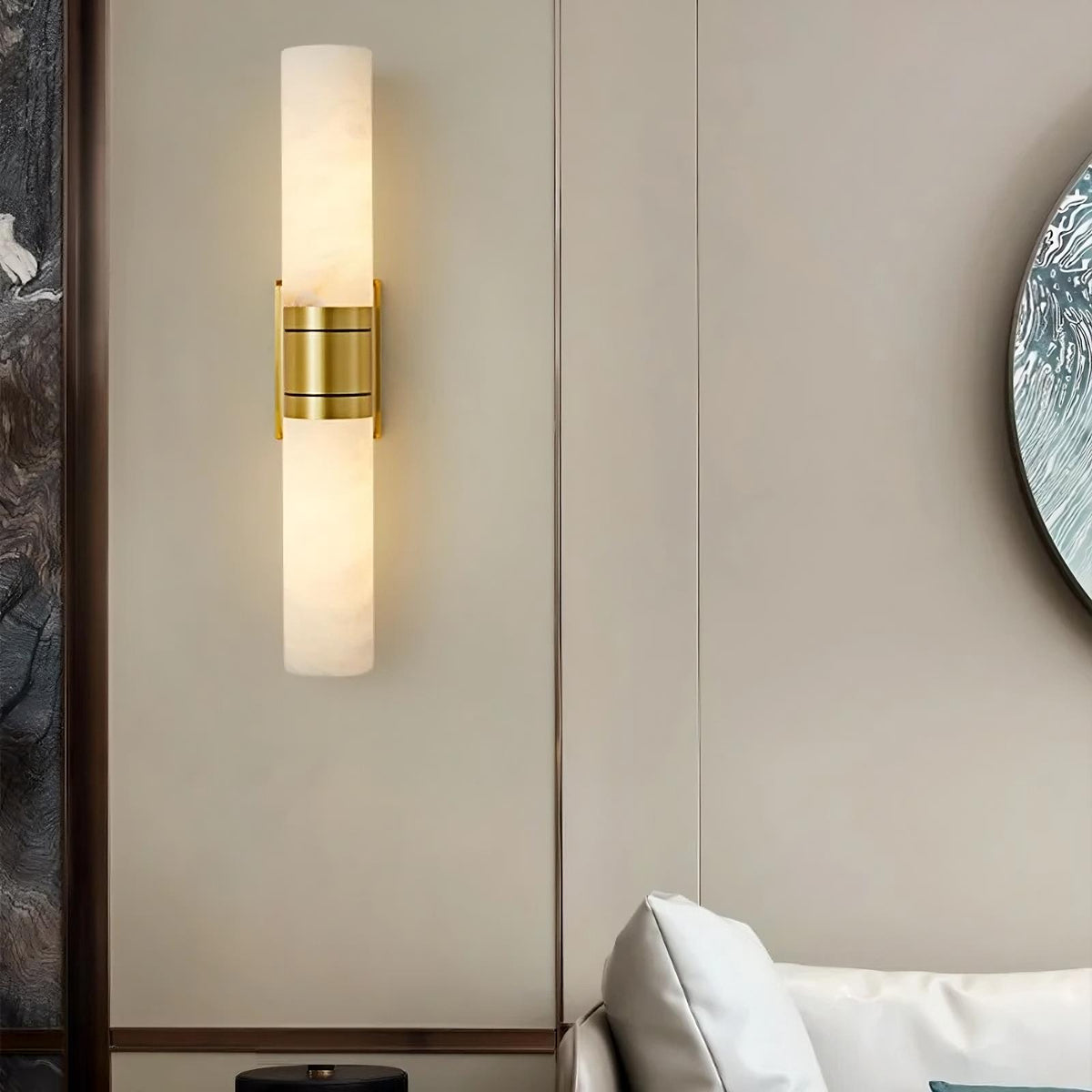 A sleek modern wall sconce with a cylindrical white diffuser and brass accents is mounted on a beige wall. This elegant Natural Marble Indoor Wall Sconce Light by Morsale.com is positioned next to a white sofa with a cushion. Part of a round, decorative mirror with unique texture is visible on the right.