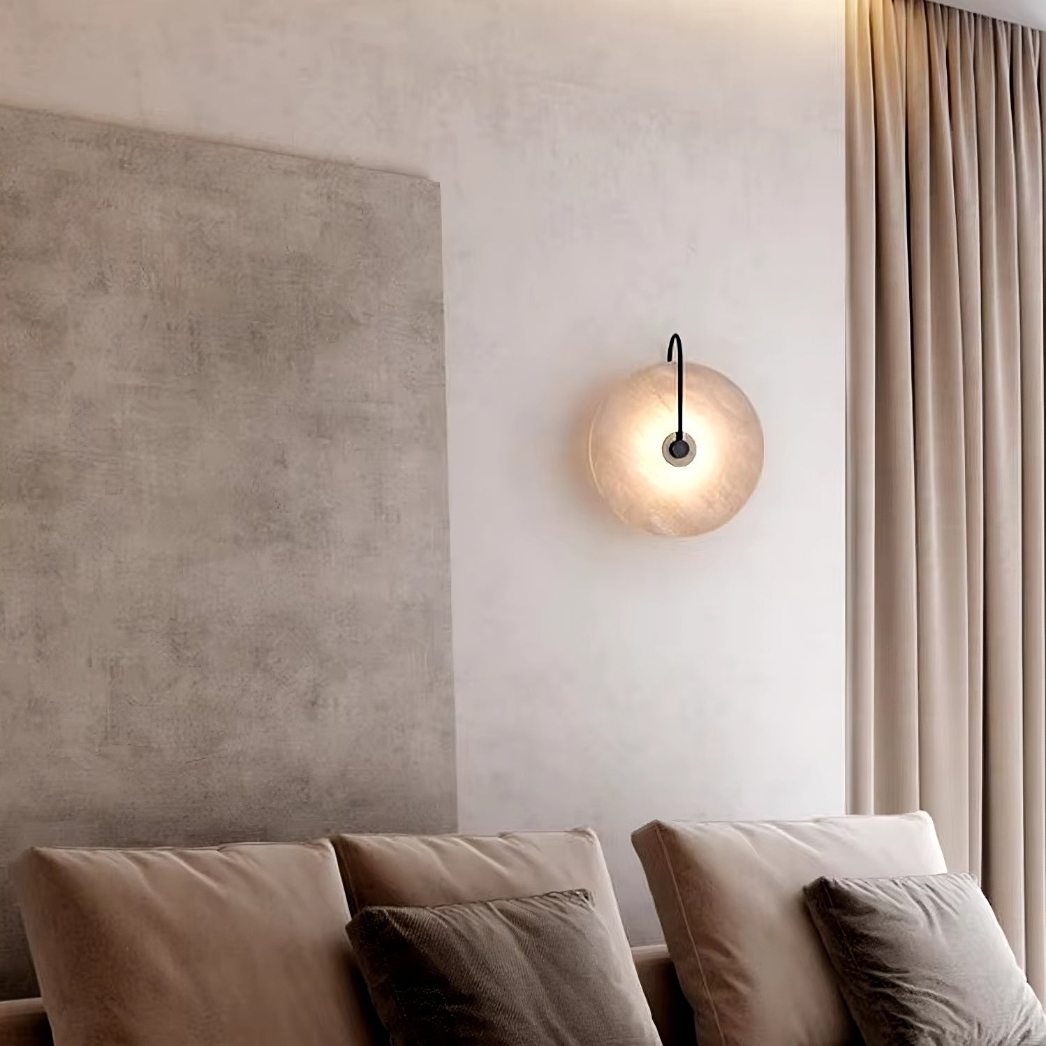 In a minimalist room, the wall is adorned with a textured panel and complemented by the Moonshade Natural Marble Sconce Light from Morsale.com, which sits beside a modern round lamp. Beige curtains elegantly frame the space, while two soft pillows are arranged on a light cushion against the wall, crafting a serene and cozy atmosphere.