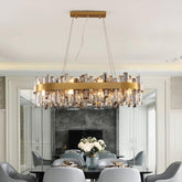 A luxurious dining room featuring the modern and ornate Asti Dining Room Chandelier by Morsale.com, adorned with clear crystals and gold accents. Below the chandelier, a stainless steel dining table set hosts a floral centerpiece. The room is tastefully decorated in neutral tones with sophisticated furniture and boasts a large mirror on one wall.