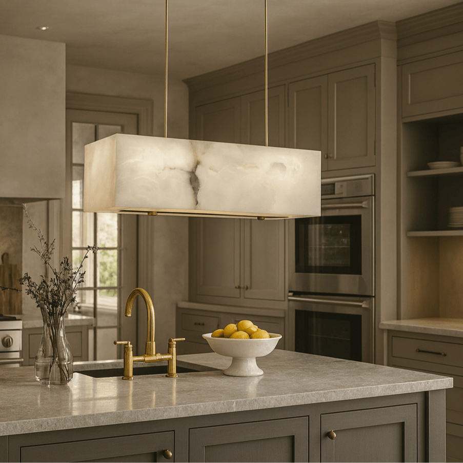 A modern kitchen features taupe cabinets, a marble island countertop, gold fixtures, a sink, lemons in a bowl, a vase with greenery, built-in stainless steel ovens, and the Leroy Marble Chandelier by Shopp578 above the island.