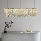 The Bacci Crystal Modern Dining Room Light Fixture 47" by Morsale.com, adorned with numerous hanging crystal-like pieces, creates a stunning cascading effect. This dimmable LED fixture is elegantly suspended above a sleek, minimalistic counter that showcases a black vase with branches and two small bird sculptures in yellow and blue against a minimalist gray backdrop.