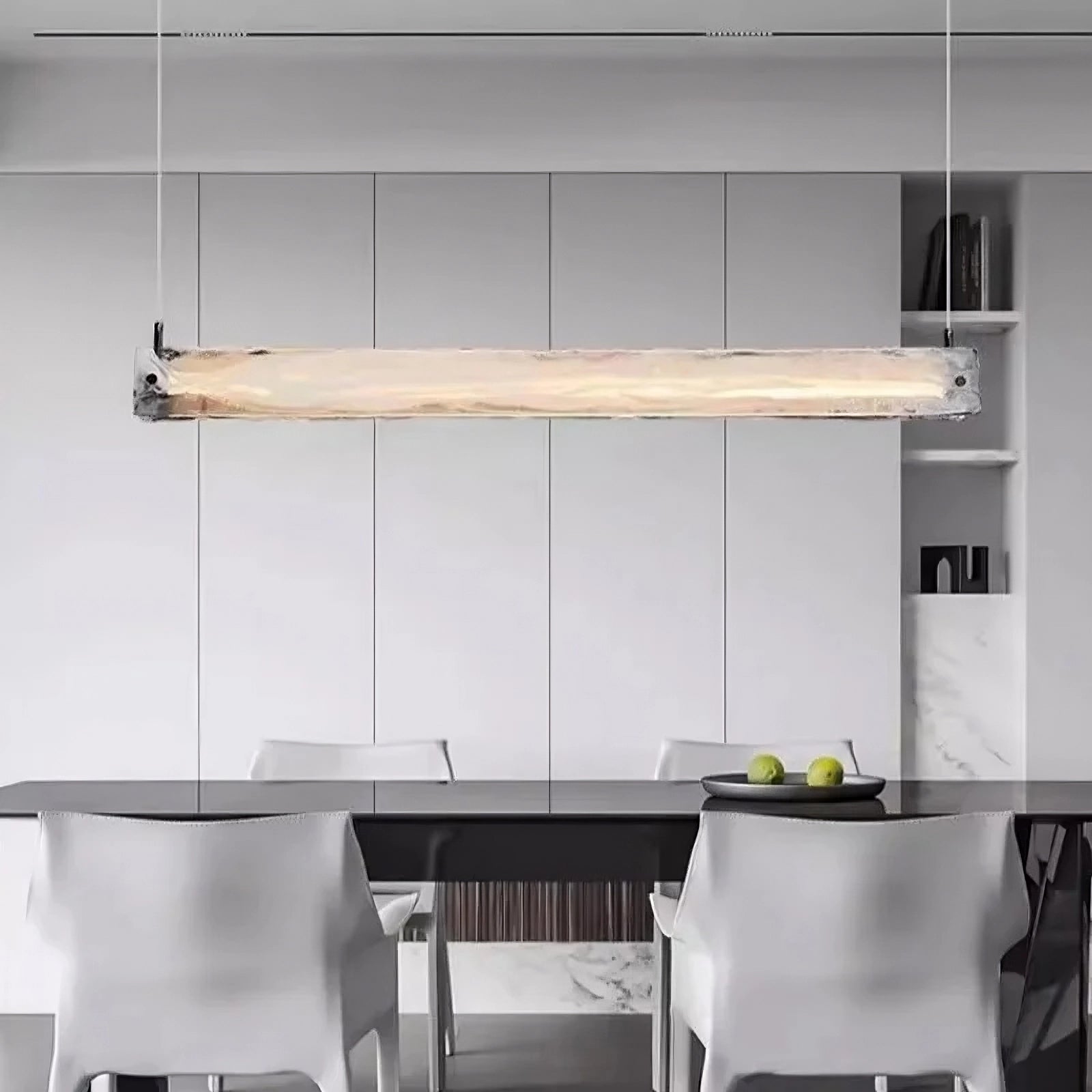 A modern luxury dining area features the Costa Dining Room Linear Suspension Chandelier by Morsale, casting a soft glow over a dark table. Four sleek white chairs enhance the scene, while white cabinetry in the background and a bowl of green apples add freshness.