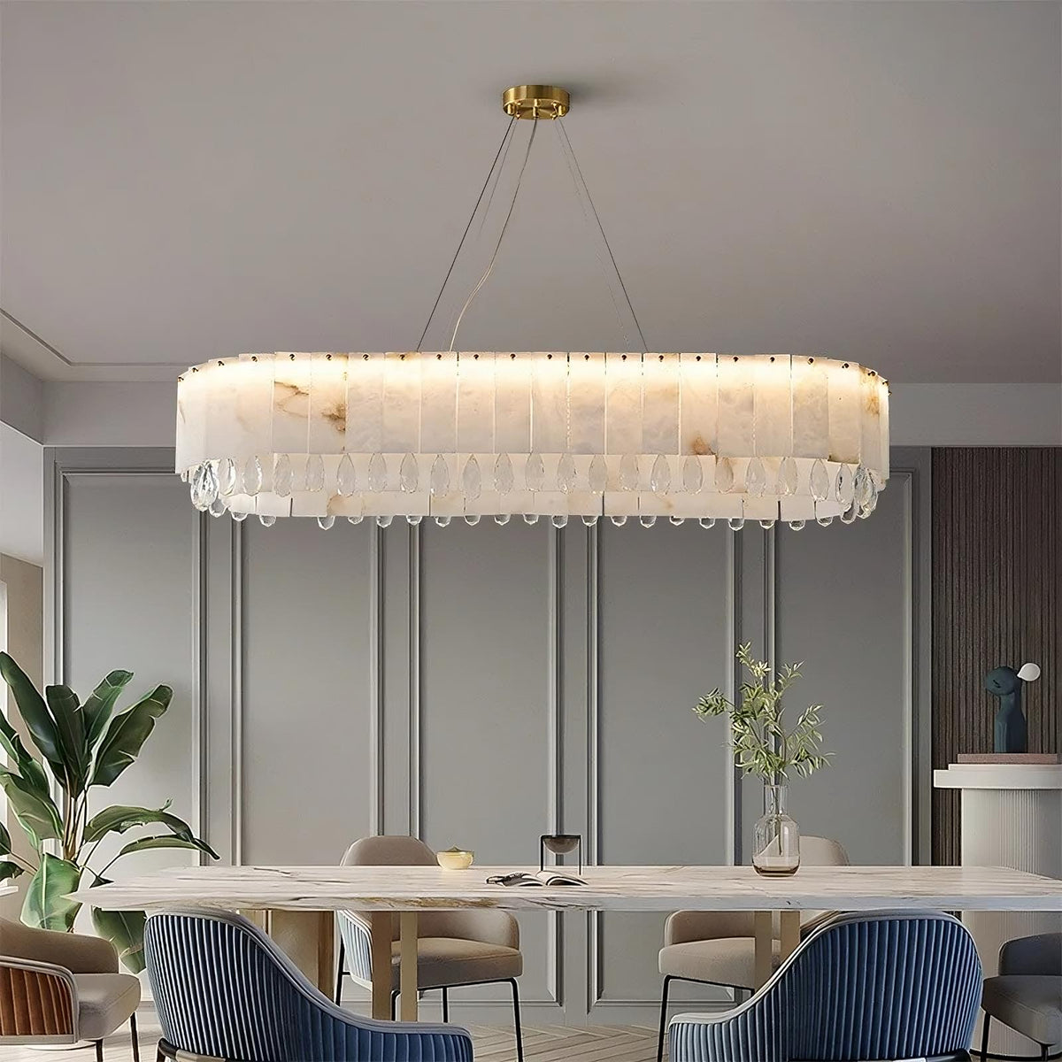 A modern dining room features a large, oval-shaped Morsale Natural Marble & Crystal Dining Room Chandelier with a translucent, decorative design and a sleek copper frame hanging from the ceiling. Below, a long dining table is surrounded by beige and blue chairs. A potted plant and vase with flowers add natural touches to the space.