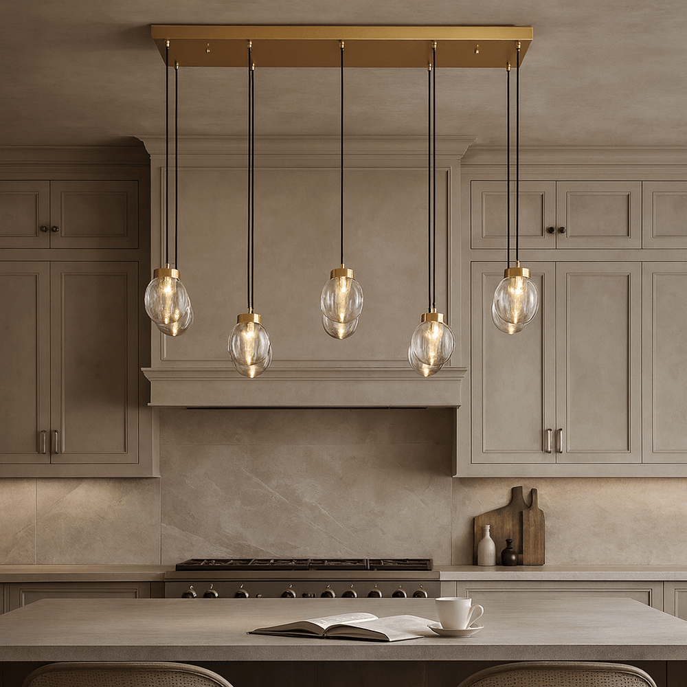 A modern kitchen with beige cabinets and backsplash features the Morsale Bossen Glass & Copper Chandelier, a gold fixture with five hanging bulbs, illuminating an island set with an open book and a cup.