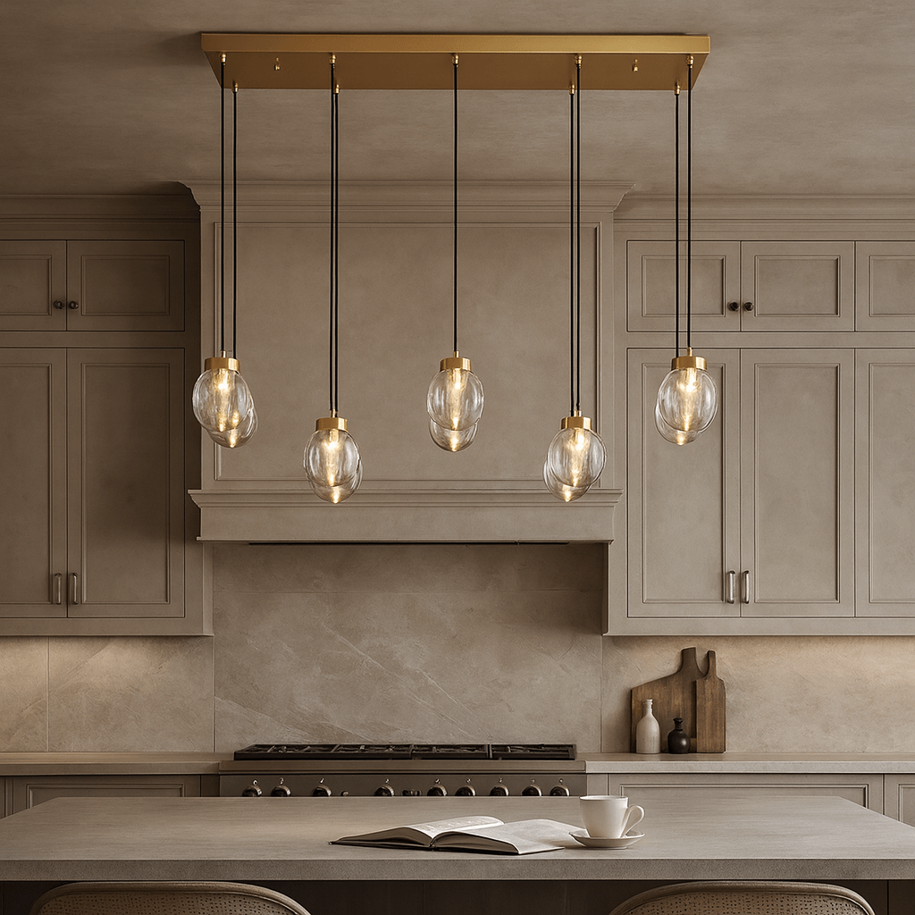 A modern kitchen with beige cabinets and backsplash features the Morsale Bossen Glass & Copper Chandelier, a gold fixture with five hanging bulbs, illuminating an island set with an open book and a cup.
