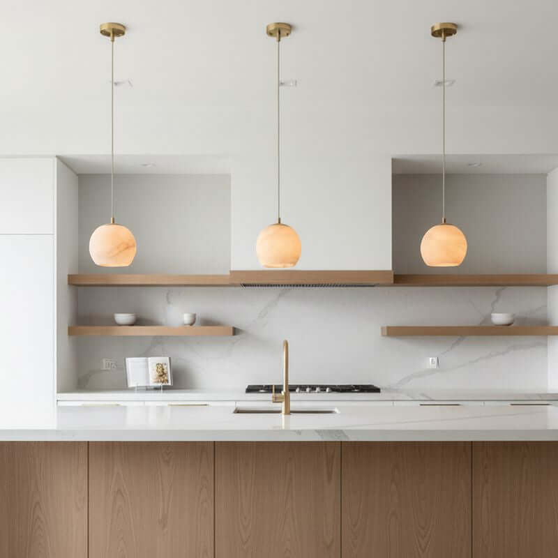 A minimalist kitchen features a marble backsplash, wooden cabinets, open shelves, and ambient light from three Auris Marble Pendant lights by Morsale.com above a white island with a gold faucet. Bowls and a book accent the space.