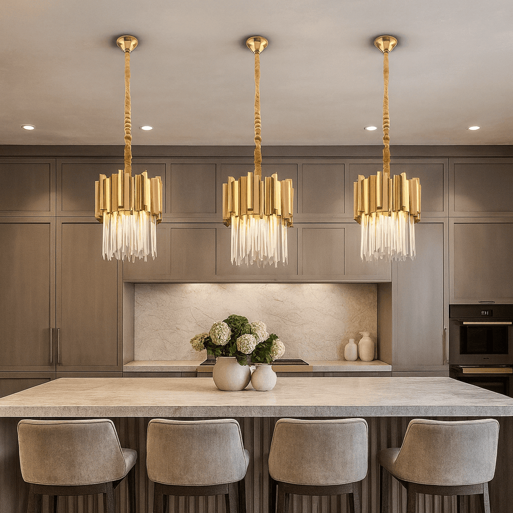 A modern kitchen features a large island illuminated by the Gio Crystal & Gold Pendant from Morsale.com, four upholstered bar stools, taupe cabinetry, built-in appliances, and a vase of white hydrangeas as a centerpiece.