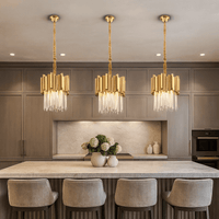 A modern kitchen features a large island illuminated by the Gio Crystal & Gold Pendant from Morsale.com, four upholstered bar stools, taupe cabinetry, built-in appliances, and a vase of white hydrangeas as a centerpiece.