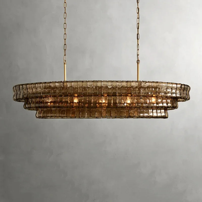 The Morsale Riviera Vintage Glass Dining Room Chandelier features elegant brownish glass tiers in an oval shape, gracefully suspended by two golden chains. It casts a warm glow against a plain light gray wall, perfect for dining room lighting.