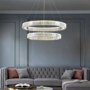 A modern living room with a tufted grey sectional sofa adorned with pink accent pillows. Above the sofa, two Roma 2-Ring Crystal Chandeliers from Morsale.com hang from the ceiling. A small round coffee table holds a book and white cup. The walls are painted grey, reflecting the room's high-quality materials.