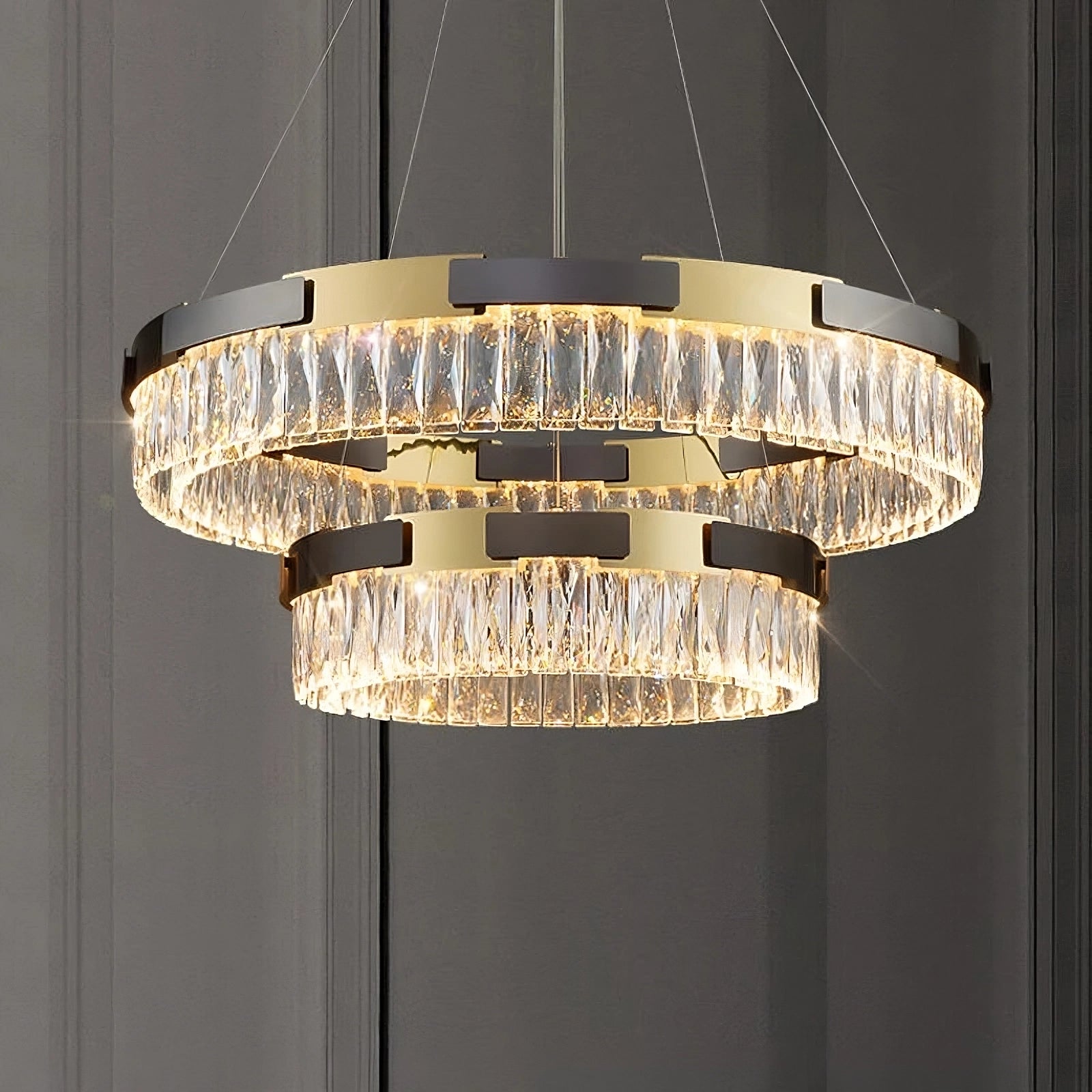 The Siena Contemporary Crystal Light Fixture by Morsale.com is a modern chandelier featuring two circular tiers, both embellished with clear crystal pendants that beautifully reflect light. The fixture boasts a metallic gold frame and is elegantly suspended by thin wires from the ceiling, set against a gray backdrop.