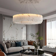 A modern living room with a large, Morsale.com Marble & Crystal Modern Chandelier hanging from the ceiling. The room features a white sectional sofa adorned with pillows, a marble coffee table with decor, and tall windows allowing natural light in. The walls have a marbled accent.