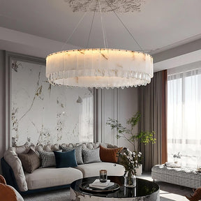 A modern living room featuring a large, elegant Modern Crystal and Spanish Marble Chandelier by Morsale.com with a translucent design, hanging from the ceiling. Below, there's a plush sectional sofa with various cushions, a round coffee table, a potted plant, and a floor-to-ceiling window with sheer curtains.