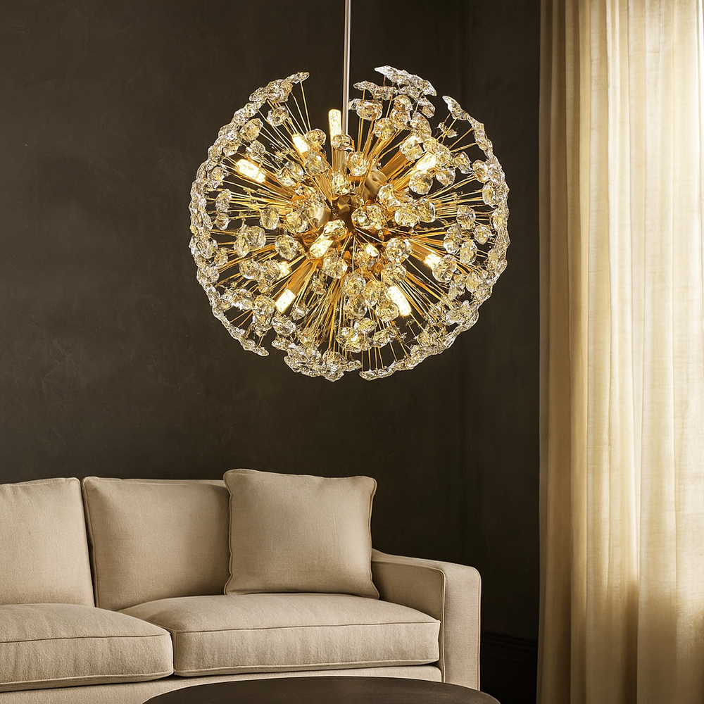 A modern living room features a beige sectional and the Aurelio Dandelion Sphere Chandelier by Morsale.com, its sparkling crystals hanging from the ceiling. Light filters through sheer beige curtains set against a dark wall.