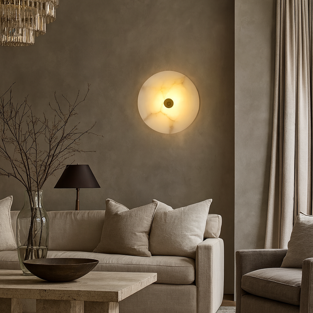 A cozy living room with a beige sofa, neutral cushions, a wooden table, bowl, lamp with a dark shade, vase with twigs, and the Hastings Marble Wall Sconce by Morsale.com illuminating the textured taupe wall.