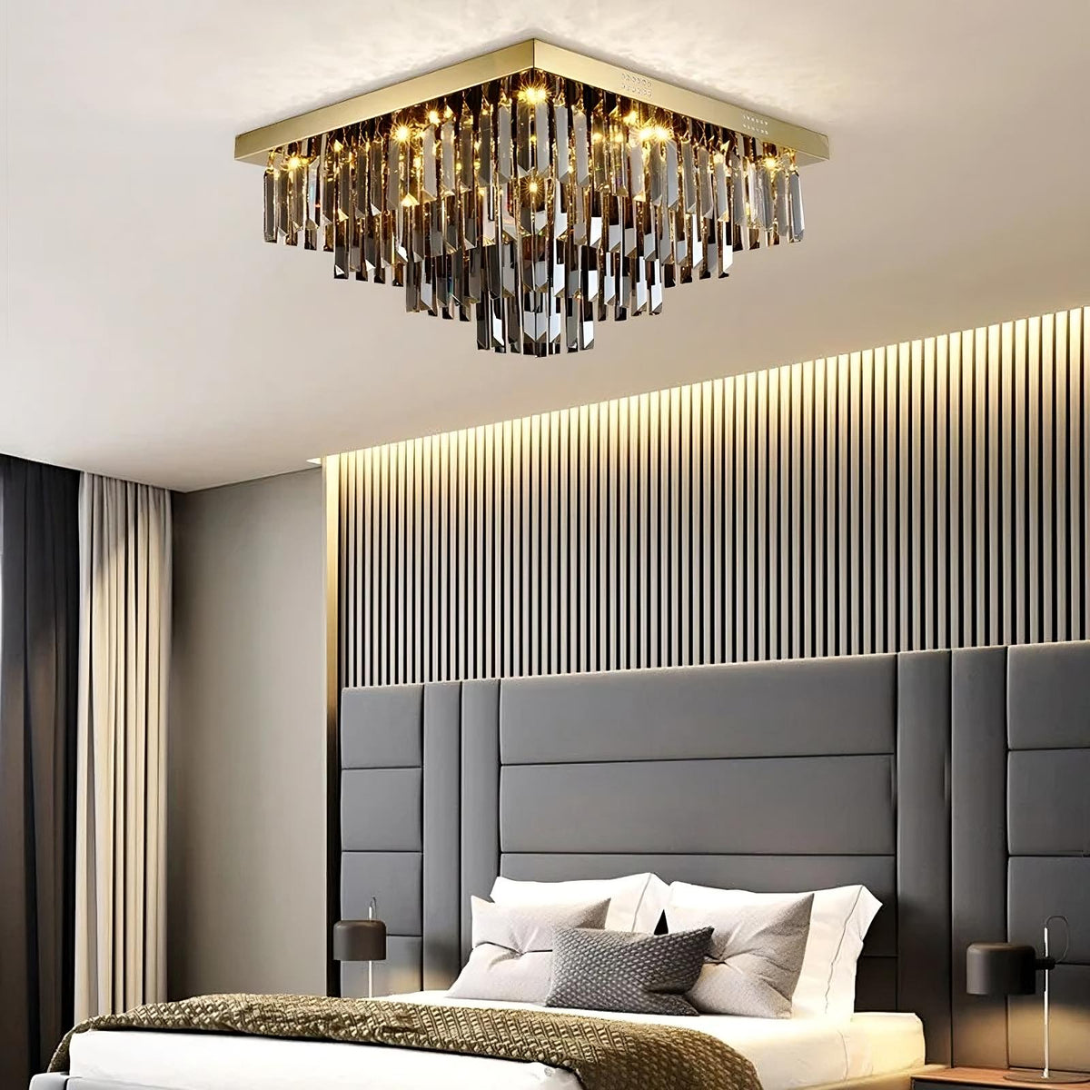 A modern bedroom features a stylish Gio Smoke Grey Crystal Ceiling Chandelier from Morsale.com, softly illuminating the space. The room has a gray padded headboard, neatly made bed with white and gray bedding, and sleek side tables with black lamps. Vertical panels adorn the wall, adding to the elegant design.