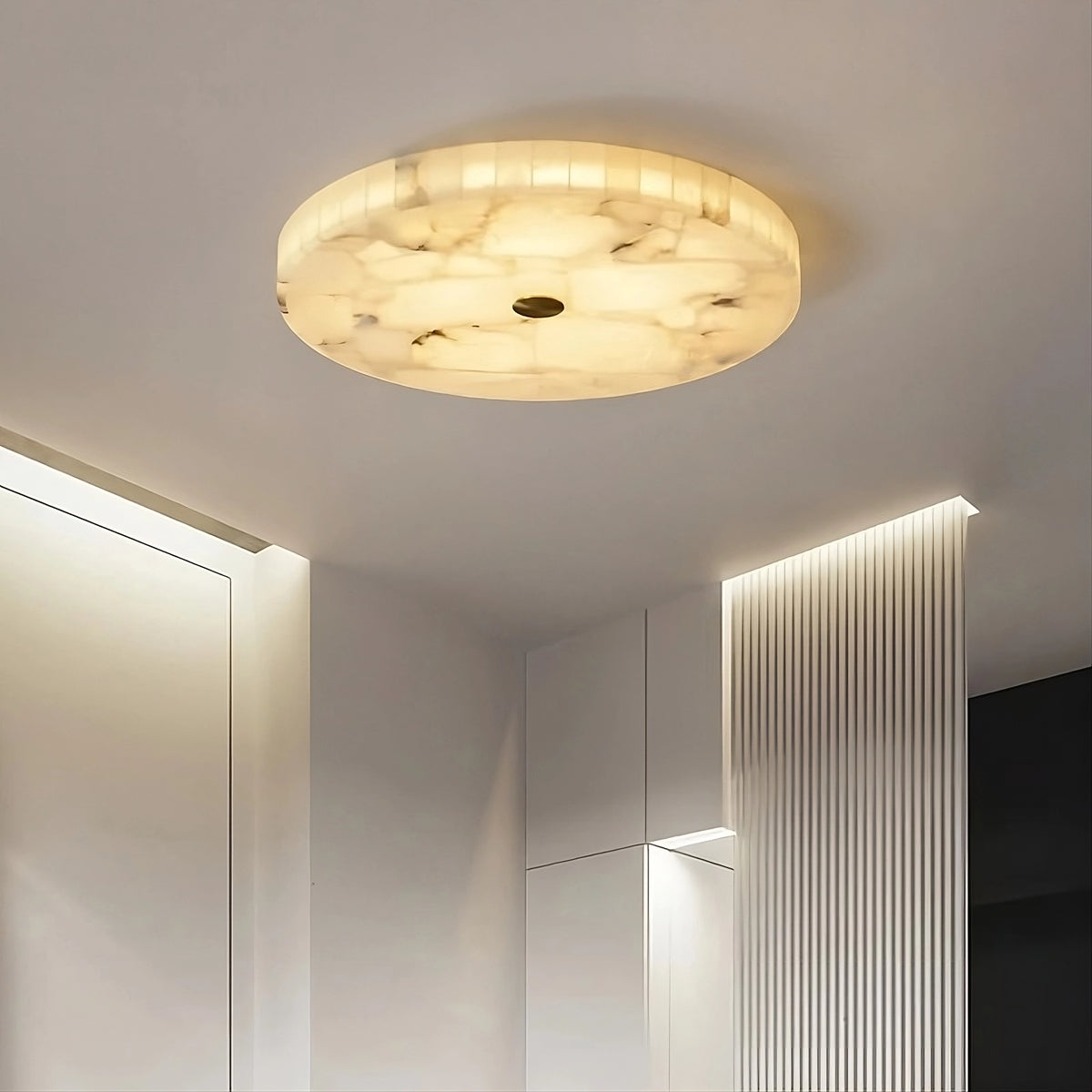 A modern room with a large Moonshade Natural Marble Ceiling Light fixture by Morsale.com that emits a warm, diffused glow. The brass-accented ceiling light has a circular design with a natural marble, translucent appearance. The room features minimalistic, sleek white cabinetry with vertical grooves and walls with clean lines.