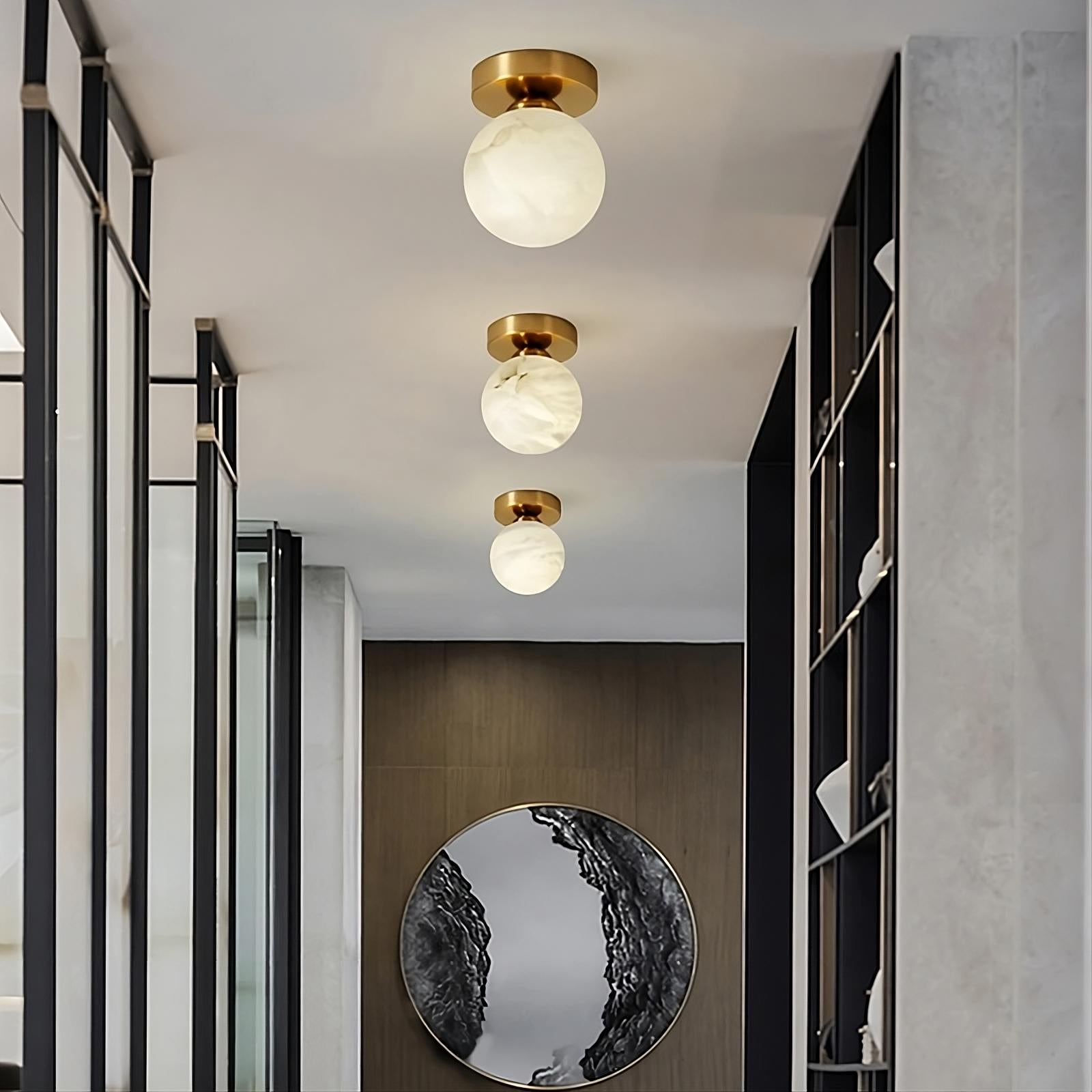 A modern hallway with three round Natural Marble Hallway Ceiling Light Fixtures by Morsale.com featuring gold bases and frosted globes. The floor has a reflective circular mirror mounted on a dark wooden panel, while the walls have metal and glass shelving units. The decor is minimalistic and elegant, enhanced by Spanish marble accents throughout.