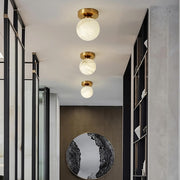 A modern hallway with three round Natural Marble Hallway Ceiling Light Fixtures by Morsale.com featuring gold bases and frosted globes. The floor has a reflective circular mirror mounted on a dark wooden panel, while the walls have metal and glass shelving units. The decor is minimalistic and elegant, enhanced by Spanish marble accents throughout.