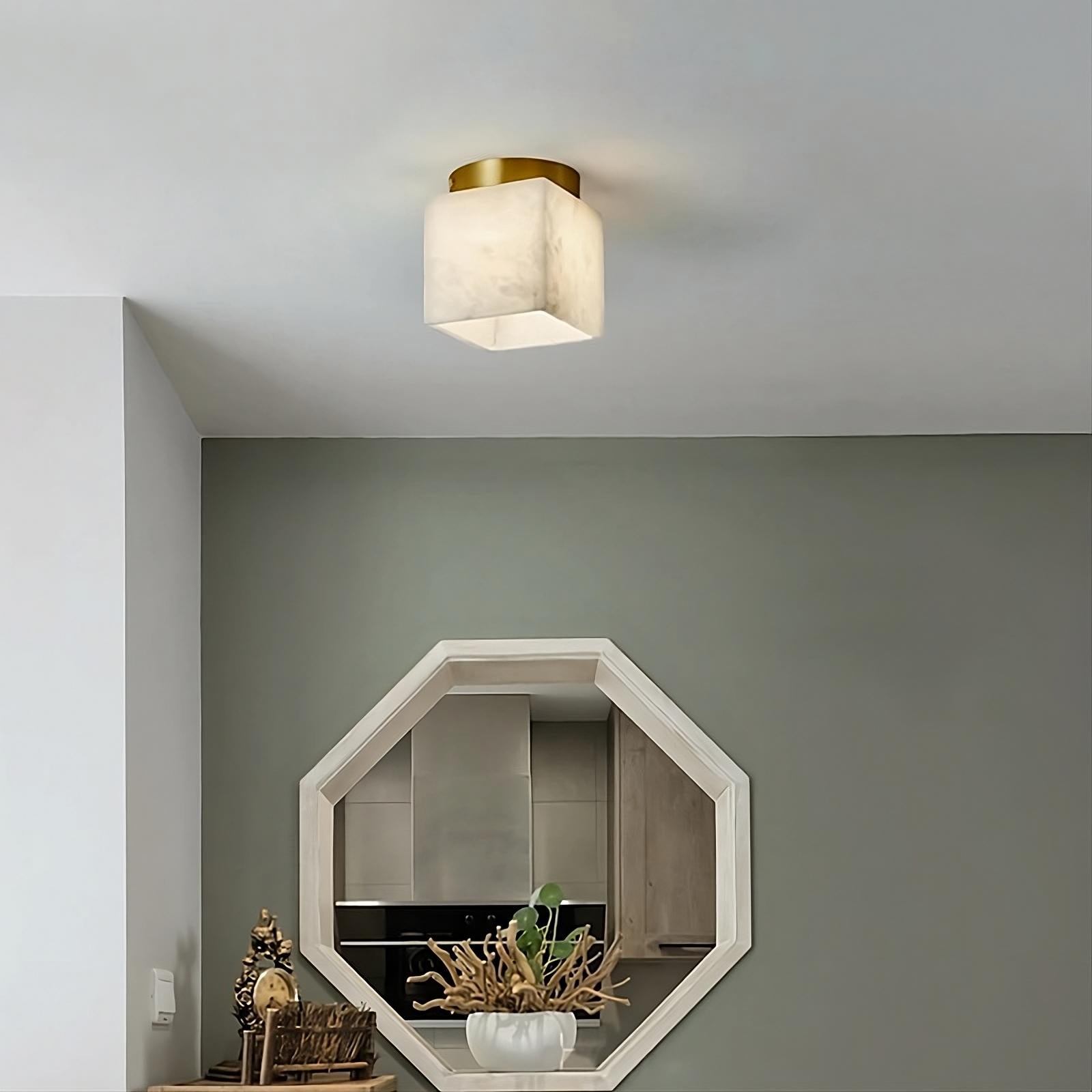 A modern room with a green and white color scheme. A Natural Marble Hallway Ceiling Light Fixture by Morsale.com is prominent. Below it is a hexagonal mirror mounted on the wall, reflecting part of the kitchen. The small table nearby, adorned with decor items, stands out against the Spanish marble backdrop.