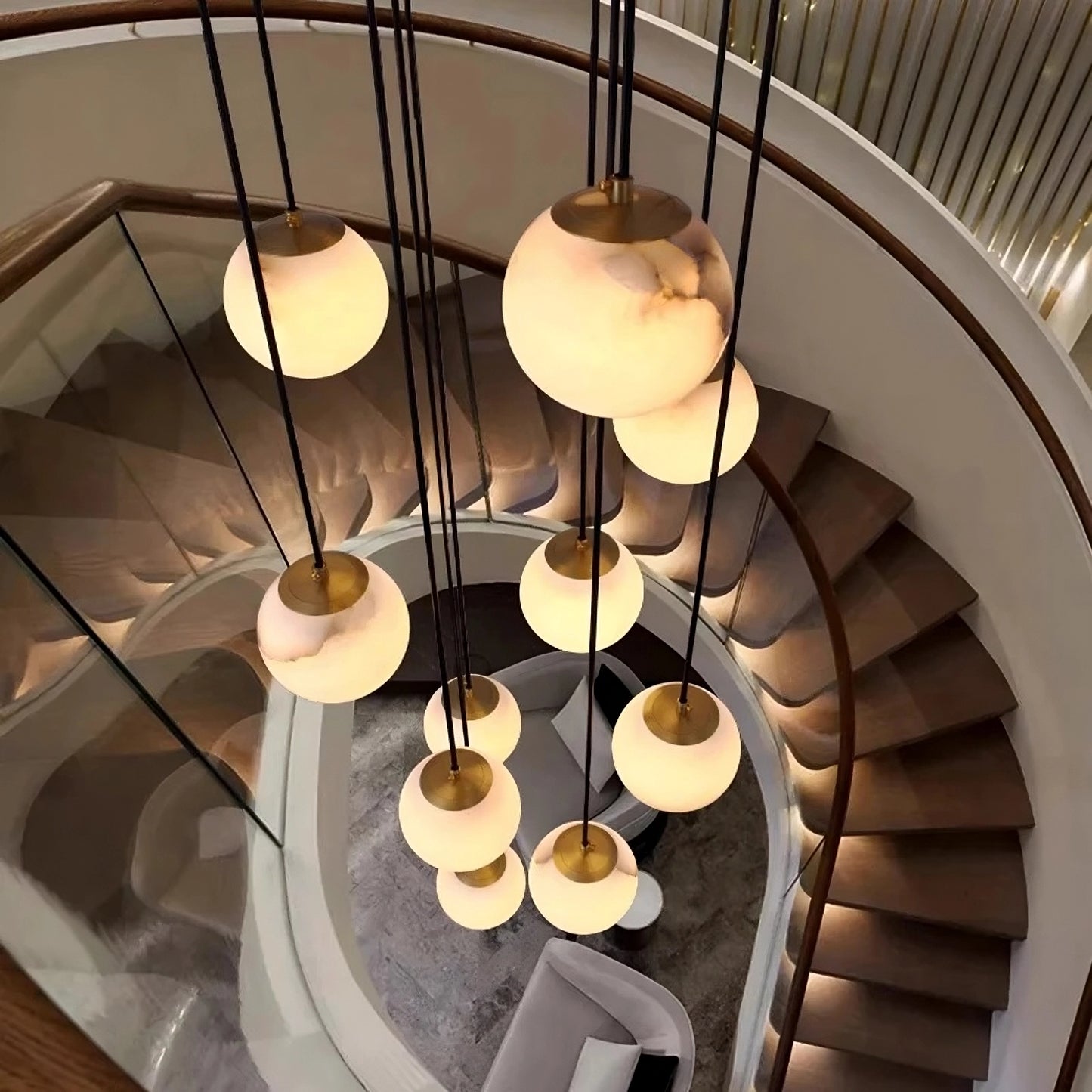A spiral staircase with wooden steps and a glass railing, viewed from above, showcases the Morsale Brighton Marble Ball Chandelier hanging in the center and illuminating the modern, elegant space below.