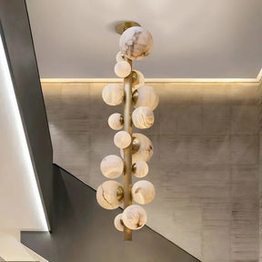 The Moonshade Luxury Marble Ball Chandelier by Morsale hangs elegantly from the ceiling, showcasing its contemporary design with spherical marble-like globes and copper accents. This vertical fixture varies in globe sizes and utilizes LED bulbs, providing an artistic flair to the minimalist interior adorned with neutral-toned walls and a staircase.