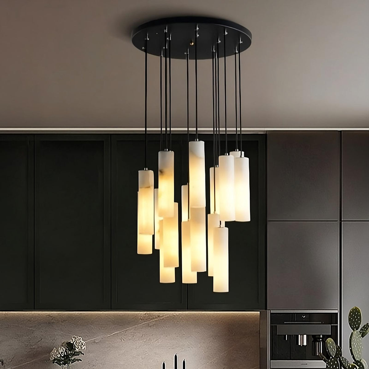 A modern 16-Pendant Marble Light Fixture by Morsale.com with multiple hanging cylindrical lights emits a warm glow. The luxurious lighting is attached to a black circular base on the ceiling of a sleek, contemporary kitchen featuring dark cabinets and built-in appliances, with some plants visible.