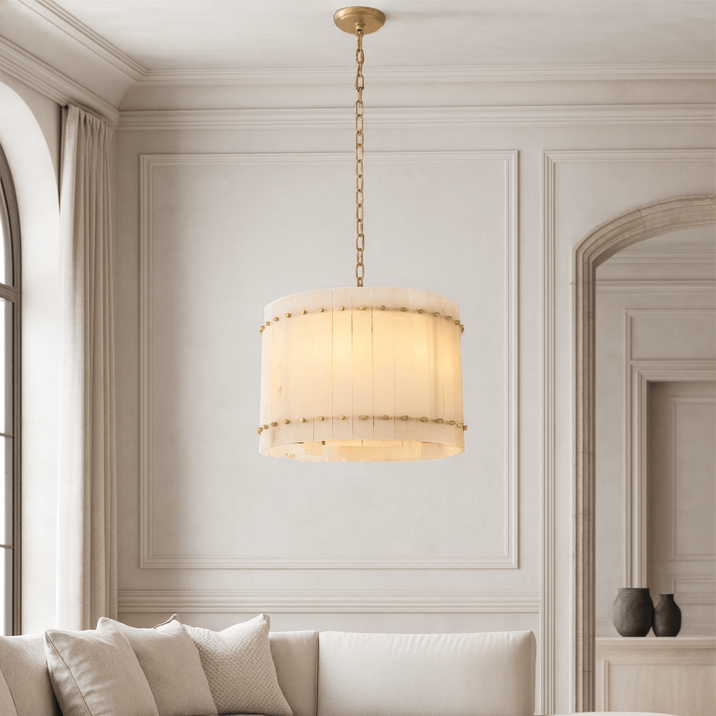 The Morsale Valmont Marble Round Chandelier 16" hangs from a gold chain in a neutral, elegant living room with paneled walls, a curved sofa, and decorative vases on the shelf.