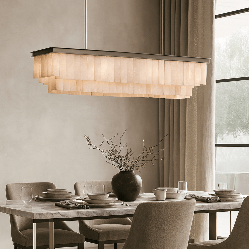 A modern dining room features a rectangular marble table, neutral upholstered chairs, and the elegant Windsor Marble Dining Chandelier by Shopp578 overhead. The table is set with plates, glasses, a dark vase with branches, and sheer-curtained windows behind.