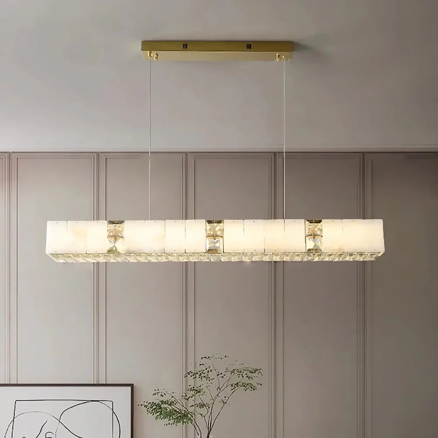 A Natural Marble & Crystal Modern Ceiling Light Fixture by Morsale.com hangs from the ceiling, featuring an elongated frosted glass shade with brass accents. The minimalist, geometric design of this luxury home décor piece complements the room's muted wall panels, abstract art, and minimalist decor.