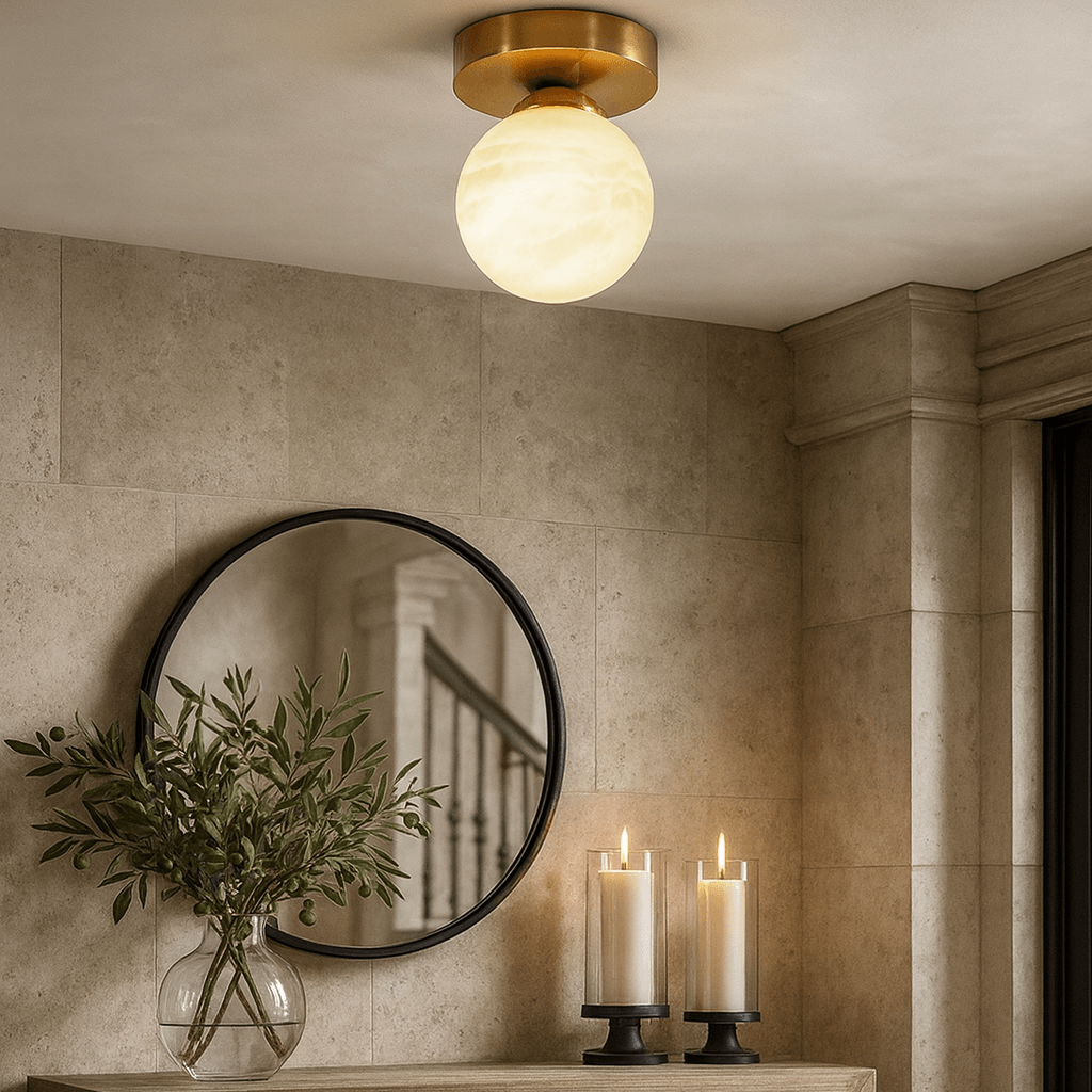 The Soria Marble Flushmount by Morsale.com, a modern round brass and glass ceiling light, is mounted on a beige tiled ceiling above a table with greenery, two lit candles, and a round mirror reflecting a staircase.