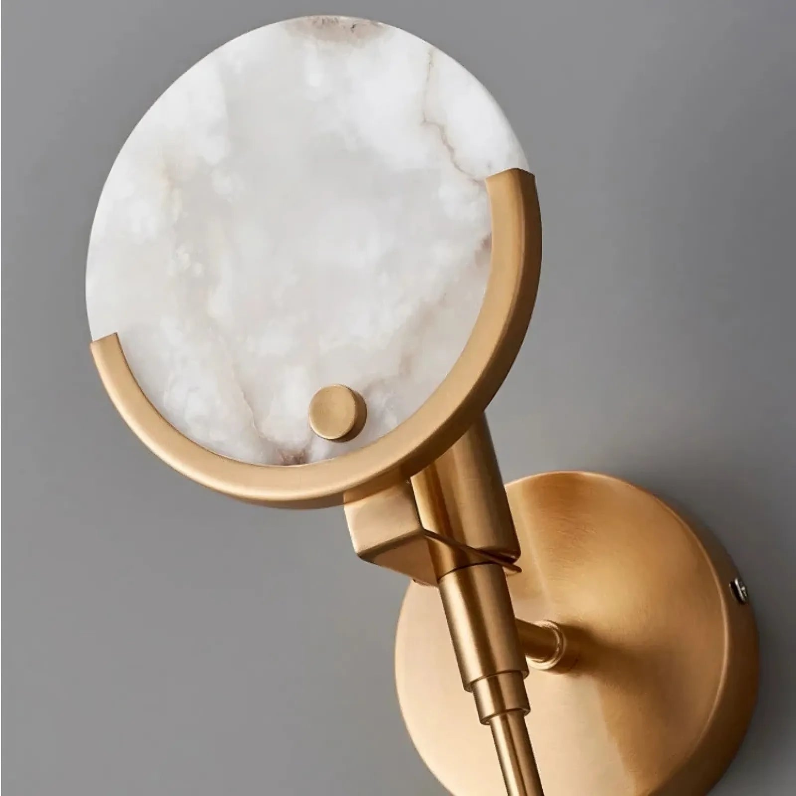 A sophisticated wall-mounted light fixture, the Natural Marble Wall Light Sconce by Morsale.com, features a circular, genuine marble shade held by an elegant brass frame. The fixture’s design combines modern and classic elements, and its smooth surfaces reflect ambient light softly, creating a warm and inviting atmosphere.