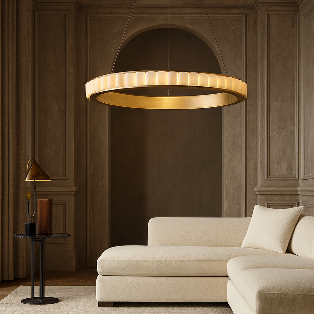 A modern living room features a cream sectional sofa, a round side table with lamp and books, and the Bigman Hampton Marble Chandelier casting warm light, all set against paneled taupe walls and floor-length curtains.