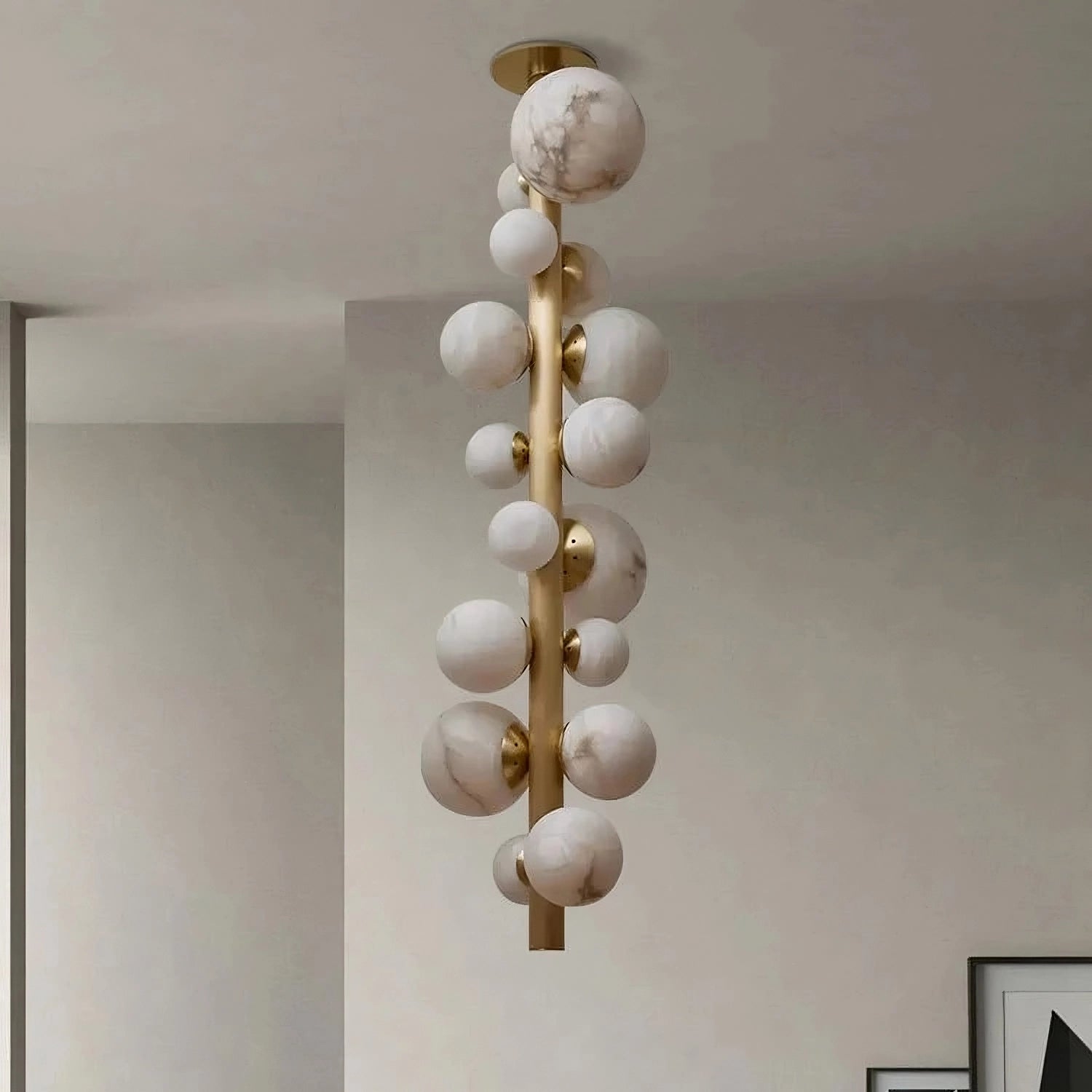 The Moonshade Luxury Marble Ball Chandelier by Morsale showcases a modern design with a vertical gold rod decorated with multiple spherical orbs that resemble white marble. This ceiling fixture, equipped with LED bulbs, stands out against a simple, light-colored ceiling and walls, offering an elegant and minimalist appearance to the room.
