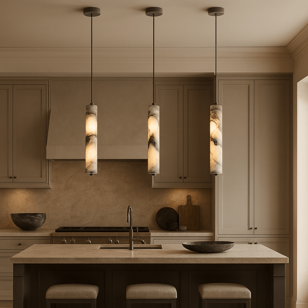 A modern kitchen with beige cabinets, a central island with a sink, three Morsale Valmont Marble Cylinder Pendant Lights overhead, two cushioned stools, and counters with decorative bowls and cutting boards accented by modern LED lighting.