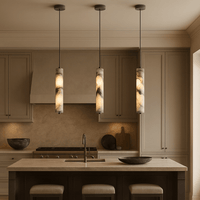 A modern kitchen with beige cabinets and countertops features three Morsale Valmont Marble Pendant Lights above an island with a built-in sink and cushioned stools, while decorative bowls and cutting boards adorn the counters.
