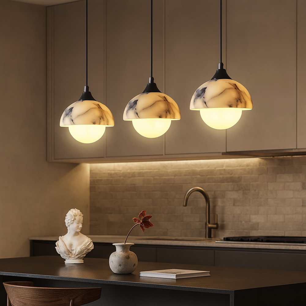 
                  
                    Three Morsale Aeris Natural Marble Pendant Lights hang above a modern kitchen island adorned with a white bust and a round vase holding a single red leaf, complemented by warm lighting and sleek neutral cabinetry.
                  
                