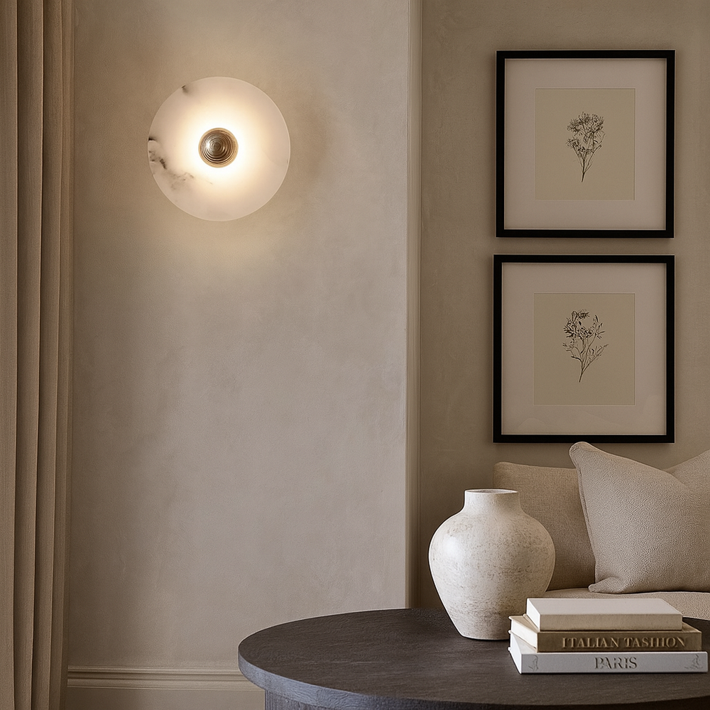
                  
                    A modern, minimalist living room corner showcases the Moonshade Marble Disc Sconce from Morsale.com, two framed botanical prints, a light-hued sofa, neutral vases, and stacked books on a round dark table.
                  
                