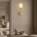 The Caldera Marble & Copper Wall Sconce by Morsale, featuring a cylindrical white shade, is mounted above a console table displaying a gold horse head and round marble bowl. Neutral-toned furniture appears in the background.