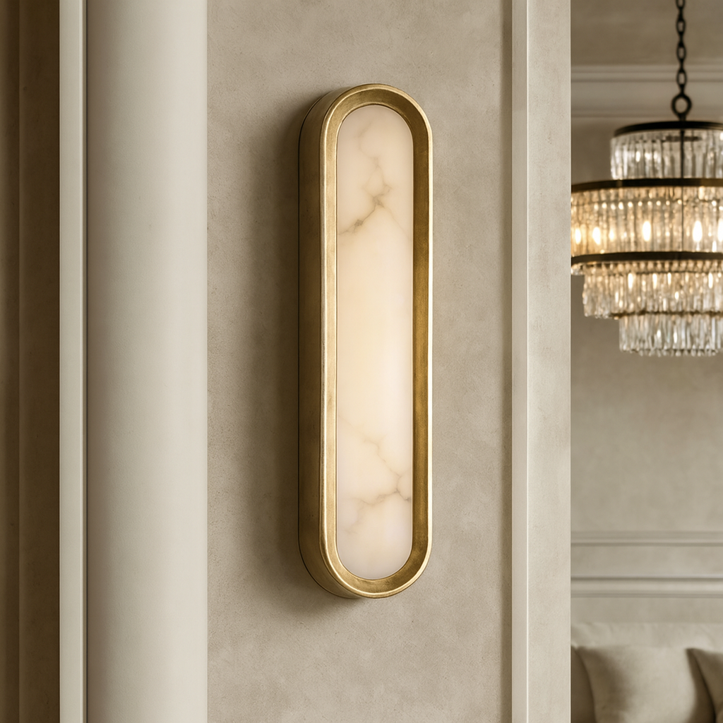 The Solari Marble & Brass Sconce by Morsale showcases a brushed brass frame and frosted marble-patterned glass on a beige wall, with a crystal chandelier elegantly illuminating the background interior.