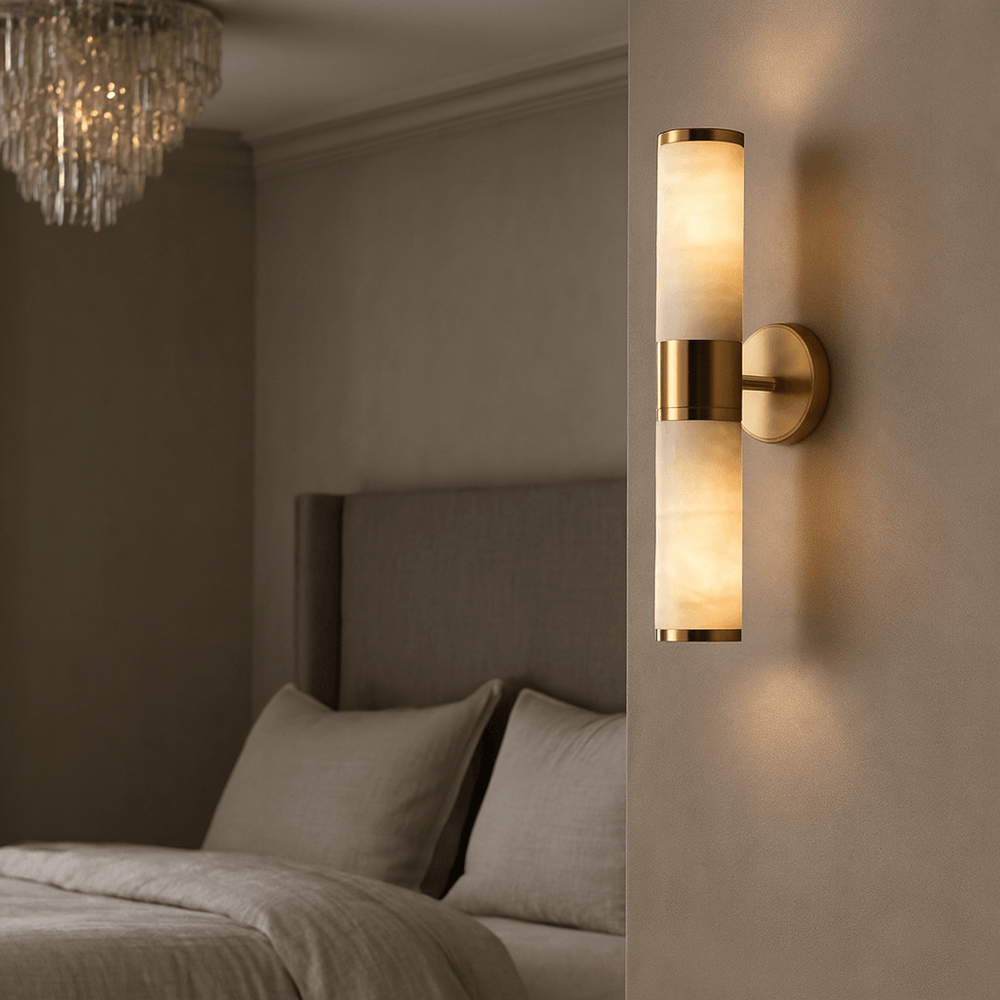 
                  
                    The Aurelia Natural Marble Wall Sconce by Morsale.com, featuring a gold finish and frosted cylindrical shade, is mounted beside a gray upholstered bed in an elegantly lit bedroom with neutral decor and a crystal chandelier.
                  
                