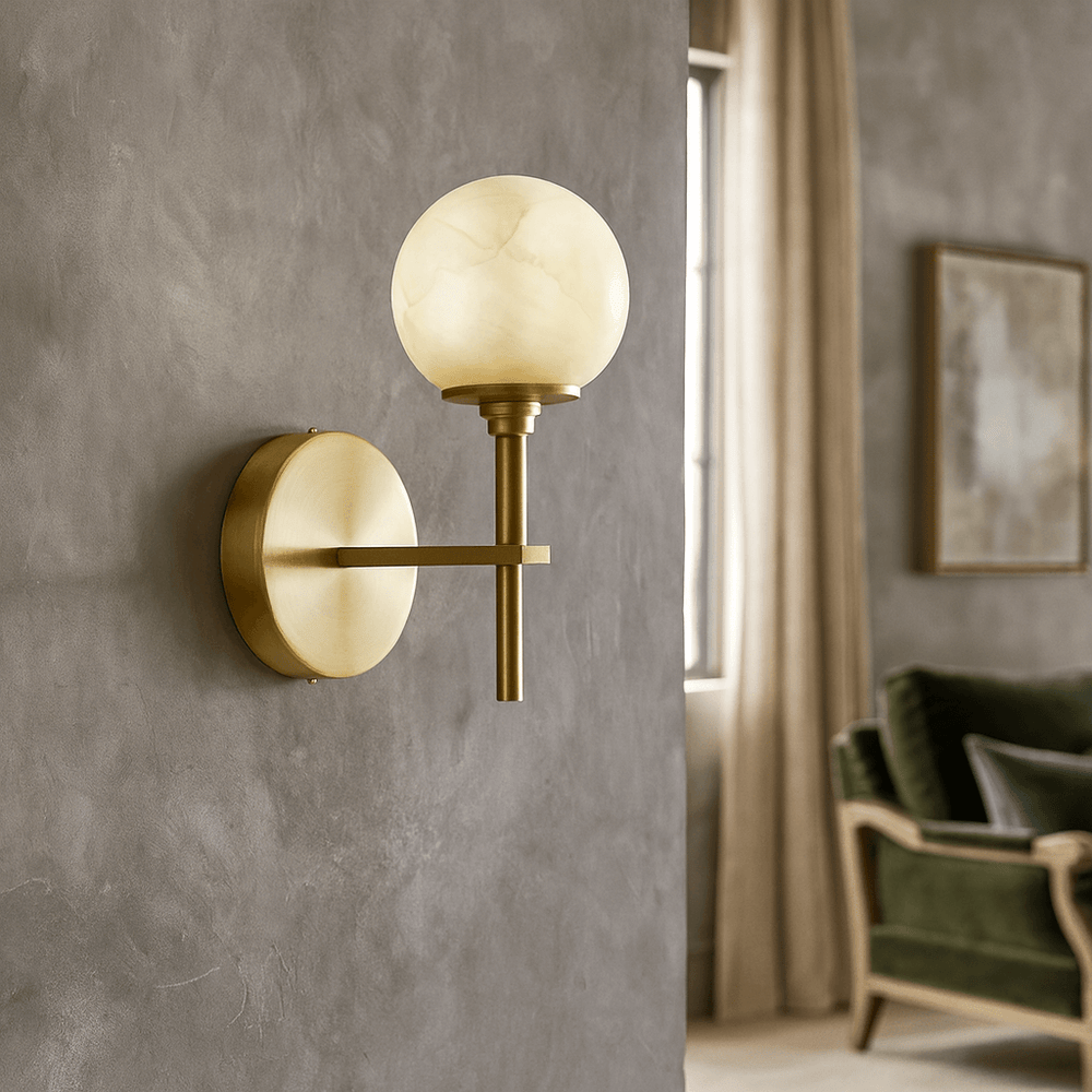 The Aria Copper & Marble Wall Sconce by Morsale, featuring a round frosted glass shade, is mounted on a gray textured wall. A green velvet chair, beige curtains, and a framed picture appear in the background.
