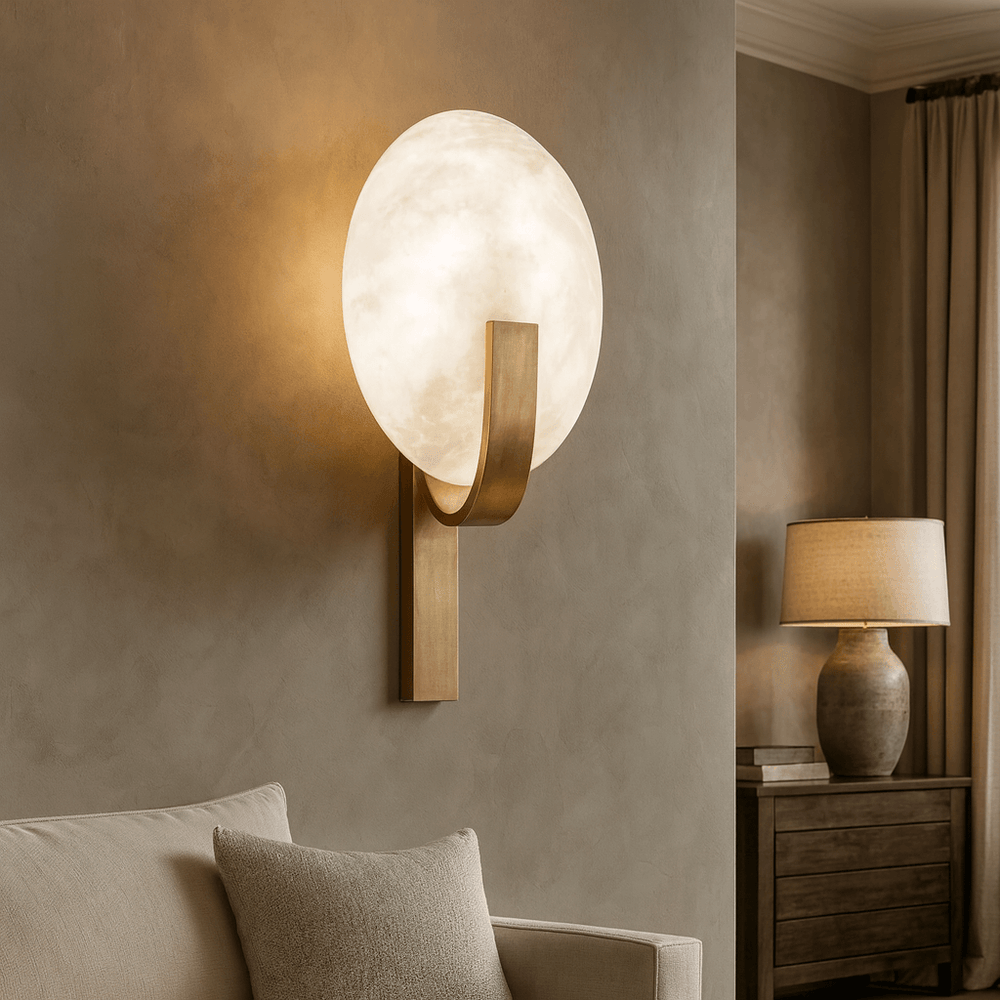 The Elara Natural Marble & Brass Wall Sconce by Morsale.com, featuring a glowing round frosted glass shade, is mounted above a beige sofa. In the background, modern luxury lighting, a table lamp, and beige curtains complete the space.