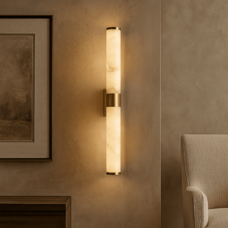 The Altura 27" Marble & Copper Sconce by Morsale features two alabaster glass tubes and copper accents, emitting warm integrated LED light, mounted on a beige wall near artwork, a fabric chair, and a wooden table.