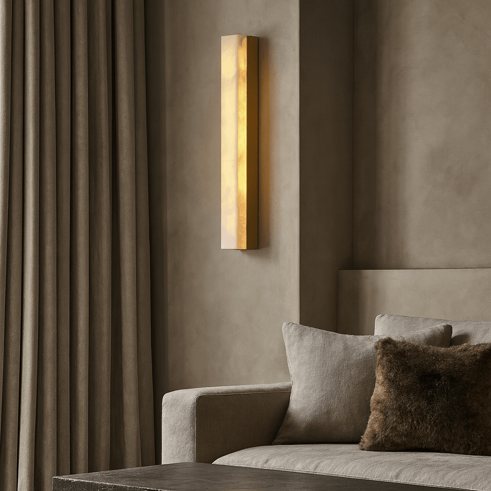 The Linden Marble Sconce by Morsale.com casts a warm glow beside a beige sofa with a brown pillow, set against textured taupe walls and floor-length curtains in this minimalist living room.