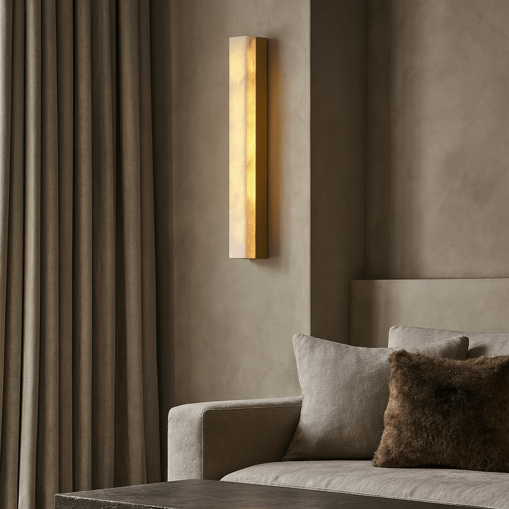 The Linden Marble Sconce by Morsale.com casts a warm glow beside a beige sofa with a brown pillow, set against textured taupe walls and floor-length curtains in this minimalist living room.