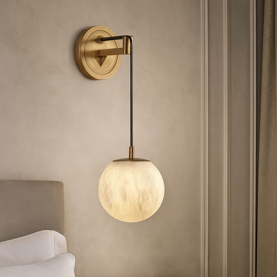 The Morsale Horrin Marble Ball Wall Sconce, featuring a round frosted glass shade and brass arm, is installed above a bedside table next to a white-bedded bed, set against a neutral wall with trim and beige curtains.