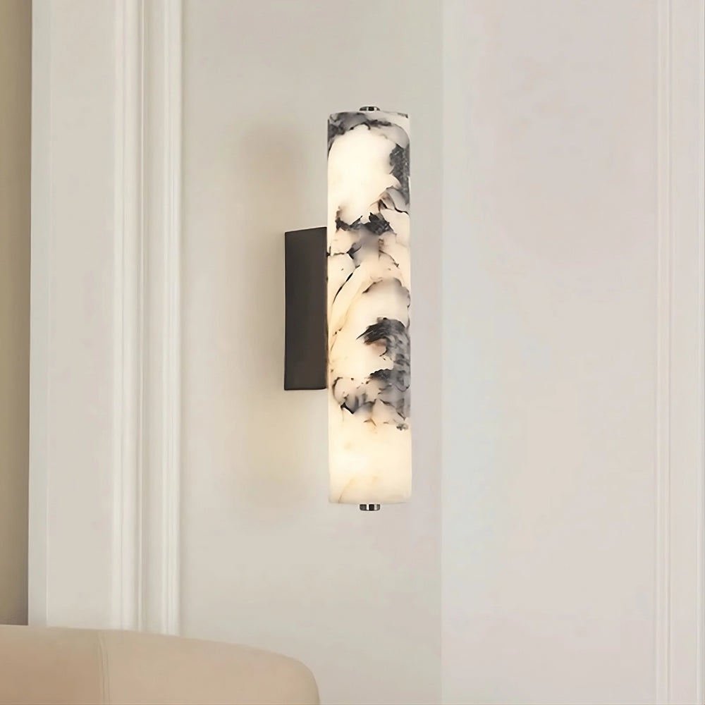 The Valmont Marble Cylinder Sconce by Morsale features a cylindrical marble-patterned shade and emits soft light. It is mounted on a light-colored wall above a beige upholstered chair.